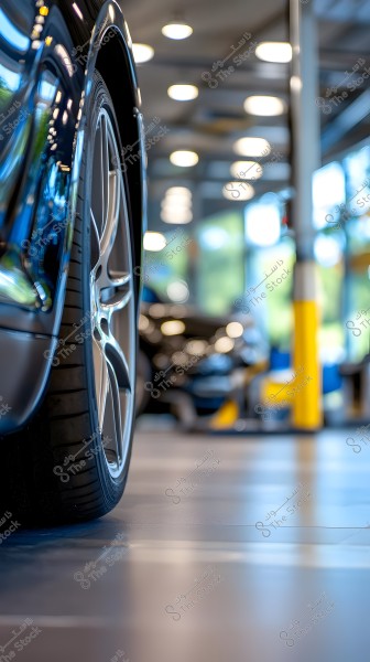 The image shows part of a modern car inside a garage. The focus is on the left front wheel with subtle reflections on the car body. The background features a yellow column and window glass revealing trees and natural light.