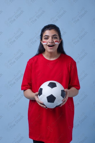 Portrait of a Saudi woman wearing a red T-shirt cheering the football team on a blue background and showing expressions of joy and enthusiasm, World Cup.