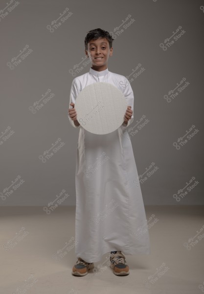 An image of a young boy wearing a white thobe standing in front of a plain gray background. The boy is holding a circular white object and smiling.