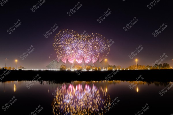 The image shows a stunning display of fireworks at night above a uniquely designed building resembling tents. The colorful reflections beautifully appear on the calm water surface in the foreground. The sky is dark, and the scene is lit by dim streetlights and the sparkling fireworks.