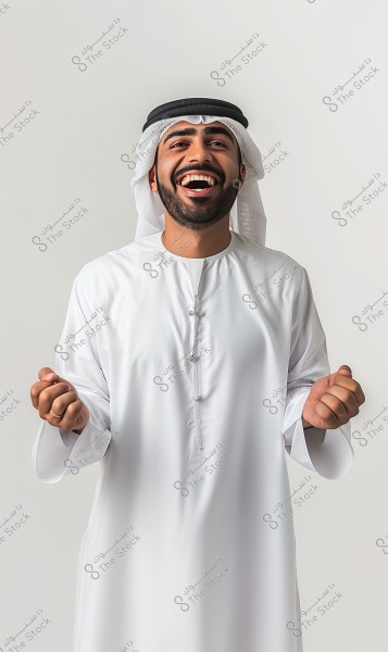 A portrait of a smiling man wearing traditional white attire, with a headscarf and a black agal. The man looks happy and is on the verge of laughing, standing against a neutral white background. The clothing suggests he is from the Gulf region, like the UAE or Saudi Arabia.