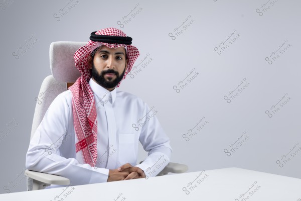 A portrait of a man sitting on an office chair. He is wearing a traditional white thobe, agal, and ghutra, suggesting he is from a Gulf country like Saudi Arabia. The background is gray, and the man is seated calmly with his hands clasped on the white table in front of him.