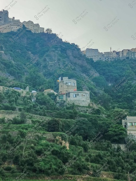 A natural landscape depicting a mountain covered with greenery, with small houses built into its slopes. The houses are integrated into the hilly terrain, surrounded by shrubs and trees. At the top, there is a cluster of large buildings on the mountain peak, creating a unique and beautiful architectural view.