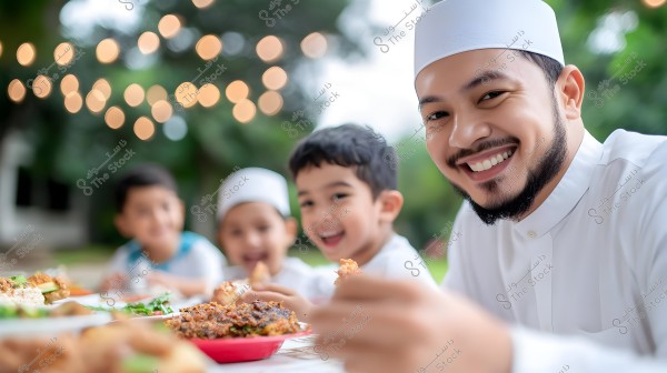 An image of a group of people sitting in an outdoor garden, with a blurred background featuring small glowing lights. In the foreground, a man wearing a white robe and a cap is smiling near a dining table with various dishes, accompanied by three children also wearing white clothes and sharing the meal.