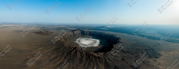 An aerial view showing a large volcanic crater in a desert area. The rim of the crater is elevated, surrounding a center that has a layer of white deposits, possibly salt or mineral evaporation. The surrounding flatlands extend with gradations of brown and gray, and the horizon stretches far into the distance under a clear blue sky.
