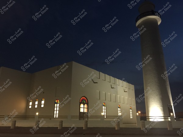 An image showing a mosque in the evening, featuring white walls and a tall illuminated minaret. There are several arched windows decorated with stained glass, adding a distinctive beauty to the design. The dark blue sky provides a calm background to the scene.