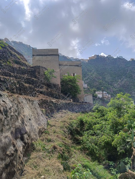 An image of a green mountainous area covered with trees and plants, interspersed with traditional stone-built houses. In the background, a range of mountains is seen under a cloudy sky with some scattered houses on the peaks.