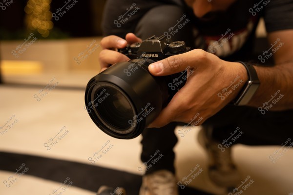 صورة ليد شخص يحمل كاميرا Fujifilm كبيرة الحجم, يلبس ساعة ذكية على معصمه. الشخص في وضعية القرفصاء, ويرتدي قميصًا داكن اللون وسروالا وجزمة. الخلفية غير واضحة مع تأثير بوكيه.