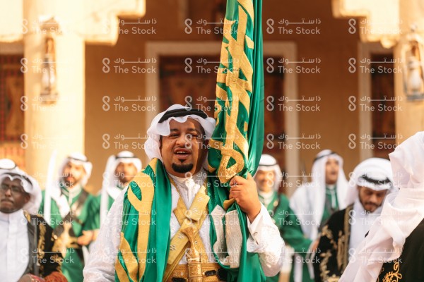 رجل سعودي يرتدي الزي التقليدي يحمل البيرق أثناء أداء رقصة العرضة التقليدية في قرية مبنية على الطراز التراثي