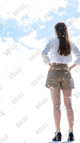 A woman stands facing the blue sky filled with clouds, wearing a white shirt, brown shorts, and black high-heeled shoes. The scene around her is filled with flying leaves or feathers.