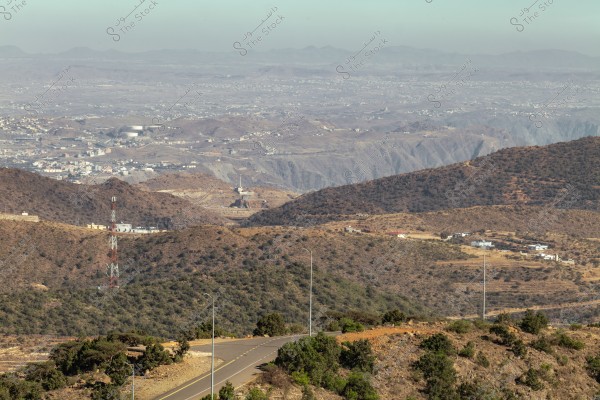 A natural landscape with rolling hills extending to the horizon. The scene features small scattered buildings. An asphalt road runs through the foreground, with a communication tower visible on the left side of the image. Trees are sparsely scattered in the foreground areas.