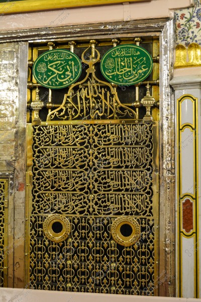 An image showing part of a shrine at Al-Masjid an-Nabawi in Medina, Saudi Arabia. It features the intricate design of a golden lattice with Arabic calligraphy inscriptions, two golden circles at the bottom, and Arabic text above the lattice with geometric and floral decorations.