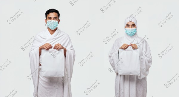 Image of a man and a woman wearing white Ihram clothing, with medical masks on their faces. The man is on the left, and the woman is on the right. Both are holding a white bag. The background is white.