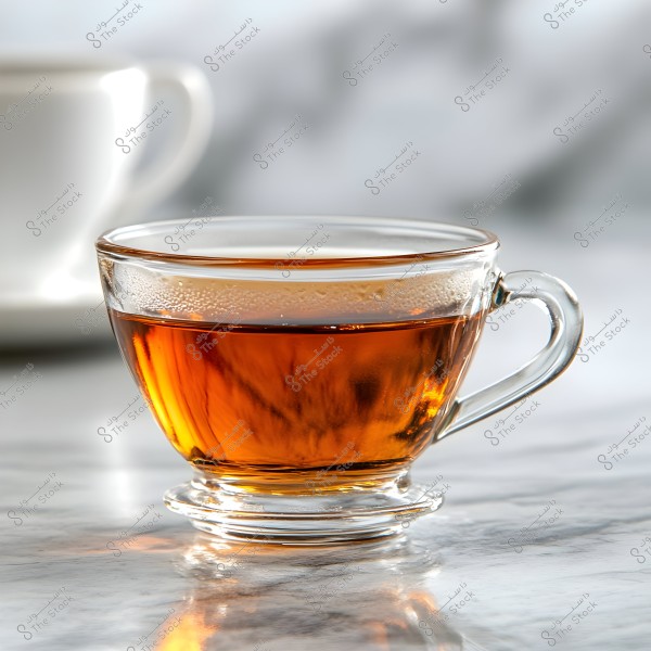 An image of a clear glass cup containing warm tea, placed on a marble surface. Steam from the tea is visible inside the cup, and the blurred background features another indistinct cup.