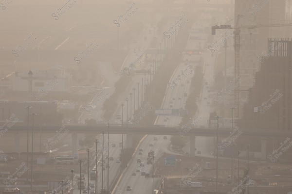 منظر لطريق سريع في منطقة حضرية خلال أجواء غبارية. عدد من السيارات تسير في كلا الاتجاهين على الطريق, الذي تصطف على جانبيه أعمدة إنارة عالية وأشجار صغيرة. تظهر مبانٍ شاهقة في الخلفية, ويوجد رافعة بناء على الجانب الأيمن. يضفي الغبار جوًا ضبابيًا على المشهد.