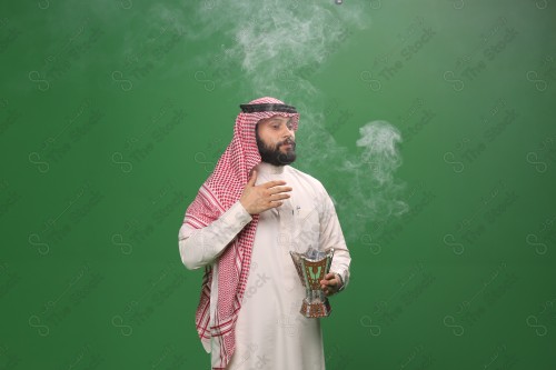 رجل سعودي يقف على خلفية خضراء يرتدي الثوب و الشماغ، يقوم بعمل حركات بيديه ، يبخر نفسه بالطيب و العود، احتفالات العيد