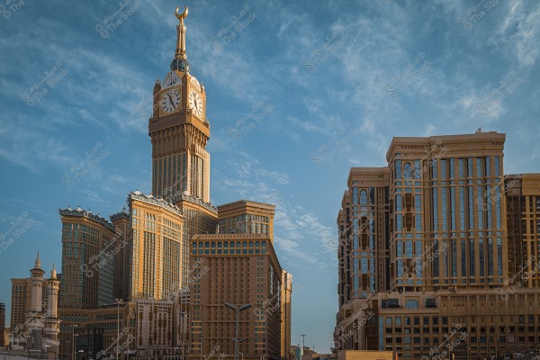Image of the Clock Tower in Mecca, known as the Royal Clock Tower. The large clock face features Islamic designs and is topped with a crescent. The building is surrounded by a complex of modern architectural towers, with a blue sky and light clouds in the background.