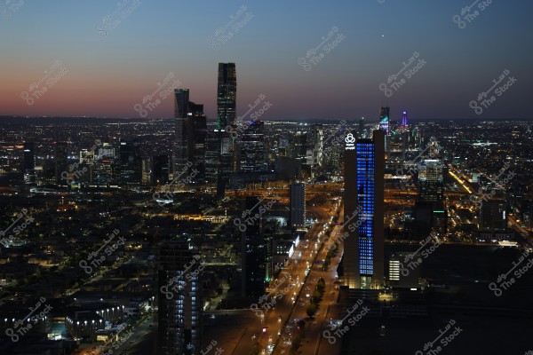 A night view of Riyadh city, featuring skyscrapers illuminated with bright lights, and cars moving along the main roads. In the background, the skyline is lit with the sparkling lights of the city.