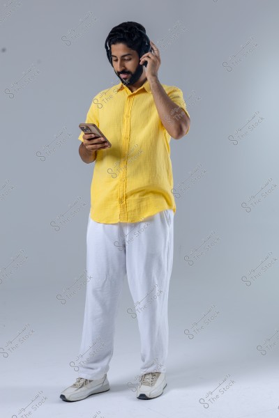 A portrait of a person standing in a studio wearing casual clothing. The person is dressed in a yellow shirt and white pants, wearing headphones. They are holding a smartphone in their right hand and appear to be listening to something. The floor and walls are light gray.