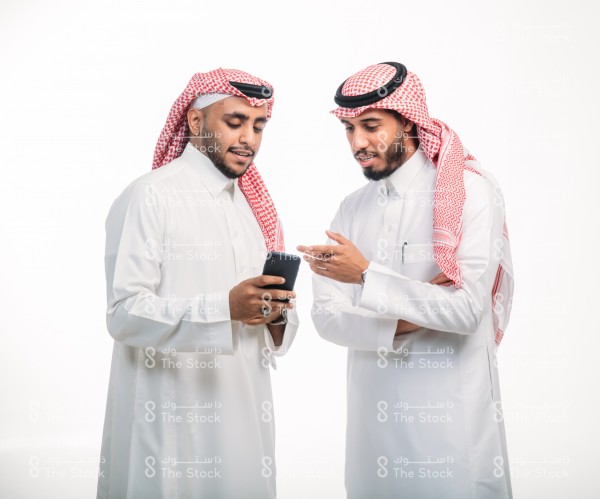 Two young Saudi men in traditional Saudi dress discussing while looking at the mobile screen