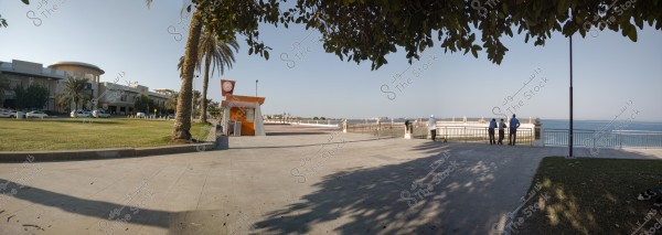 The image depicts a waterfront view in Jeddah, Saudi Arabia. On the left, there is a modern building with palm trees and parked cars in front. In the center, there is a small orange kiosk with a slanted roof. A group of people stands by a fence overlooking the calm blue sea, under the shade of lush trees. The sky is clear and bright with no clouds.