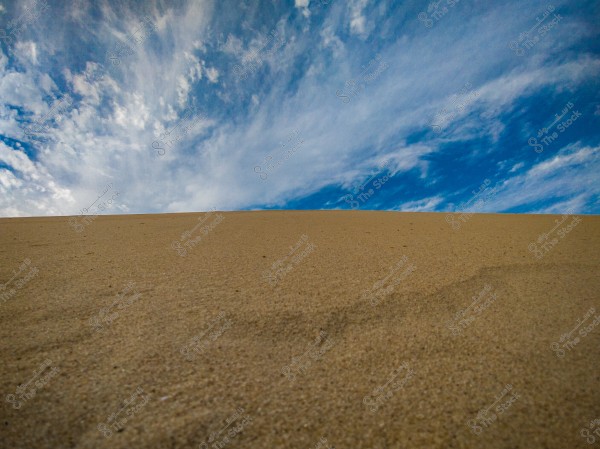 A natural scene showing vast sand dunes extending toward the horizon under a blue sky with scattered white clouds. The golden color of the sand stands out in the middle of the image, while the clouds add a dramatic touch to the sky.