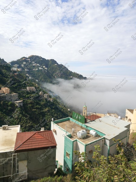 مشهد طبيعي لمنطقة جبلية تغطيها الخضرة في السعودية. تظهر في الصورة منازل مع أسطح مسطحة وخزانات مياه, محاطة بغيوم كثيفة وشجار خضراء كثيفة. خلفية المشهد الجبلي مغطاة بالغيوم البيضاء والسحب التي تضفي أجواء هادئة وسلمية.