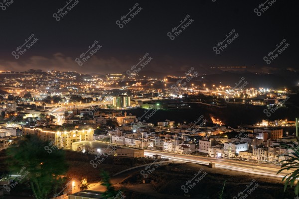 Night view of a city illuminated with lights, showing residential buildings and lit streets. Lights spread across the hills in the background, with some clouds in the sky. The highway in the foreground shows light trails from passing cars.