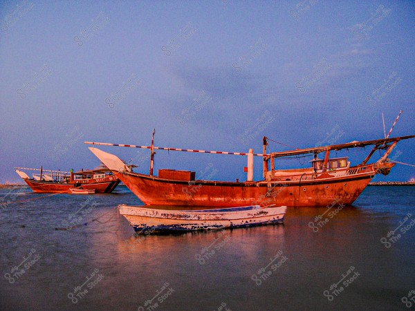 An image of traditional wooden boats anchored on the sea under a clear blue sky at sunset. The boats are large and made of wood, resembling the traditional design found in the Arabian Gulf region. The water reflects the warm colors of the boats.