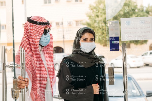 رجل سعودي يرتدي الزي السعودي التقليدي والكمامة ونظارة شمسية يفتح باب المتجر بينما تدخل امرأة سعودية ترتدي العباءة والكمامة