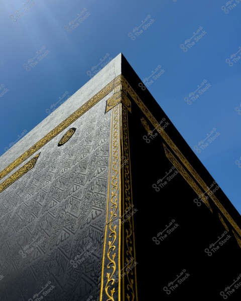 An image of the corner of the Kaaba in Mecca, showing the black cloth covering adorned with golden threads and decorative inscriptions including Quranic verses. The clear blue sky is visible in the background.