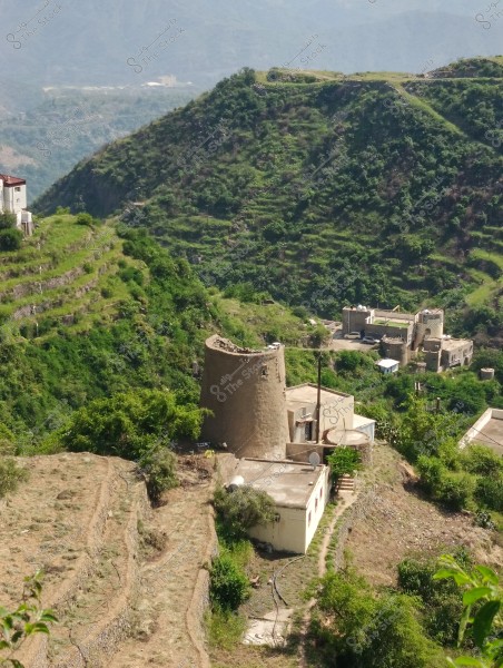 A picturesque view of a mountainous village in a rural area, featuring green hills covered with grass and trees. In the center of the image, there is an old circular tower made of stone and mud next to a small white house. Terraced fields are carved into the hills. Other houses are scattered around the area. The background includes a range of mountains under a clear sky.