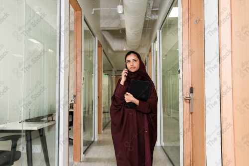 Saudi woman wearing an abaya stands in the meeting room in front of the glass front, holding a tablet ، during the day