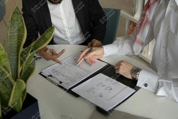 Two individuals sit around a round white table reviewing documents on a black clipboard. One is wearing a dark suit and the other is wearing a white thobe and a red checked headscarf, suggesting they may be from a Gulf country. They use pens to point at parts of the documents. A green plant in a pot is beside them.