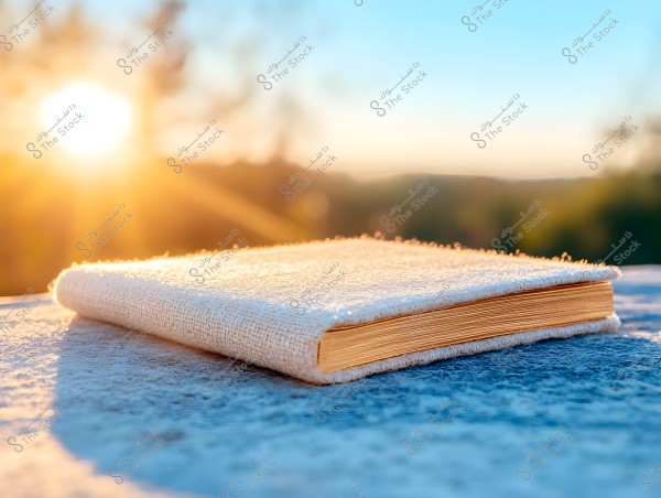 A closed book with a textured fabric cover rests on a bluish, marble-like surface, with a bright, glowing sun setting in the background, creating a warm and shimmering atmosphere. The sky is blue with blurred green trees in the background.