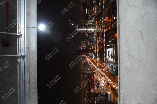 An image of a vibrant city at night, where lights illuminate the streets and buildings. Tall buildings are beautifully lit, and there is noticeable traffic on the roads. The scene appears to be taken from a high vantage point, possibly from inside a building under construction seen on the left side of the image.