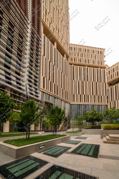 The image shows a modern building facade with a geometric architectural design, featuring prominent vertical lines and building materials of brick and glass. There are green areas and planted trees on the main pavement surrounding the building, with tiled pathways covered with glass. The sky is a light blue background, adding a touch of freshness and modernity to the scene.