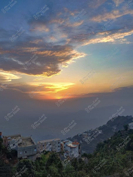 صورة لمشهد غروب الشمس فوق مجموعة من المباني على سفح الجبل. تبدو السماء ملونة بتدرجات البرتقالي والأزرق, مما يضفي جمالية خاصة على المنظر. المنازل متعددة الطوابق وتحيط بها الأشجار, وتقع في منطقة جبلية خلابة.من محافظة فيفاء بمنطقة جازان جنوب المملكة العربية السعودية