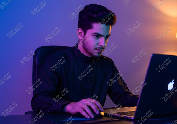 A young man seated on a chair wearing a dark shirt works on a laptop at a desk in a dimly lit environment with blue and orange lighting. He has a serious and focused expression, suggesting work or study.