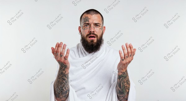 Image of a man with a beard and tattoos on his face and hands, wearing a white wrap, which resembles ihram clothing. His hands are positioned in a gesture that suggests meditation or prayer. The background is white.