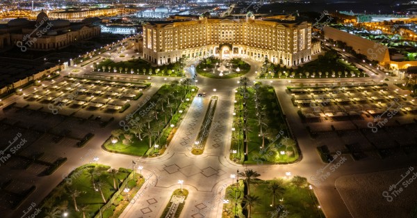 An aerial view showing a large and elegant building lit with lights, likely a hotel or a luxury palace, surrounded by well-organized gardens and illuminated fountains. The surrounding streets are organized with rows of trees and night lighting, creating an enchanting atmosphere. The city is visible in the background, illuminated with night lights.