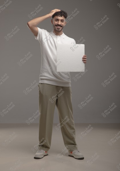 A portrait of a smiling man standing in a studio, wearing a white polo shirt, beige trousers, and white sneakers. The man is holding a rectangular piece of styrofoam in one hand while placing his other hand on his head.
