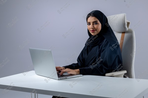صورة لامرأة ترتدي عباءة سوداء مع حجاب, جالسة على كرسي مريح خلف مكتب أبيض. تعمل على حاسوب محمول فضي اللون. الخلفية بسيطة وذات لون رمادي.