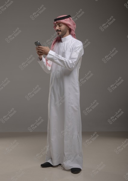 Image of a man wearing a white thobe and a red checkered headscarf with a black agal, standing and using a mobile phone with a smile on his face. The picture has a simple grey background and is a full-body portrait.