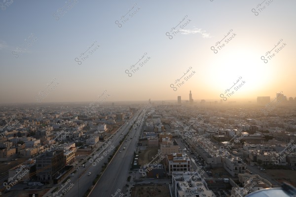 An aerial view of a city under the bright sun, showing a long road stretching toward the horizon. Buildings are situated on both sides of the road, with skyscrapers visible in the distant background. The city is bathed in the golden light of sunset, with a clear sky and a few scattered clouds.