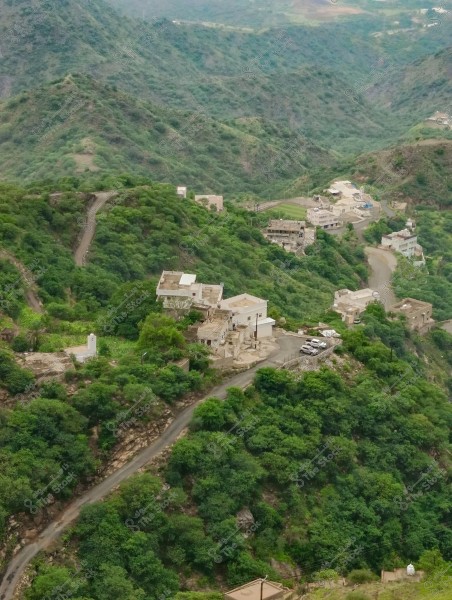 An image of a lush, green mountainous area featuring several houses and buildings scattered atop the hills. A narrow road winds through the mountains, passing by the buildings. The area is densely covered with vegetation and trees, offering a sense of tranquility and scenic natural beauty.