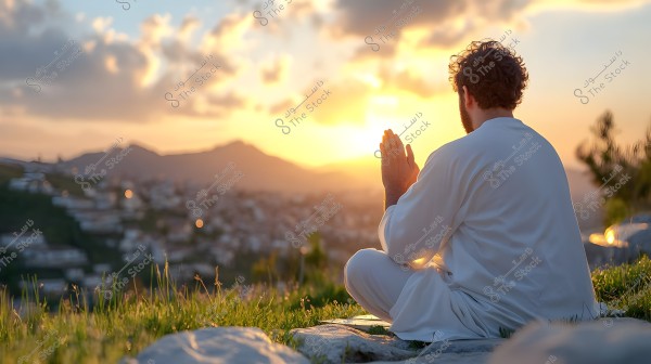 A man sitting on the ground wearing a white thobe, raising his hands in prayer at sunset. A city with mountains is visible in the background, bathed in the serene colors of the dawn sky.