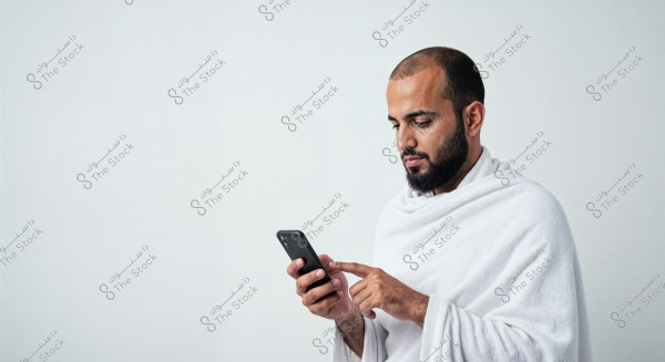 A portrait of a man wearing traditional white Ihram clothing, holding a mobile phone and looking at it intently. The background is white, and the image is in a portrait style. The origin might be Saudi Arabia or other countries where Ihram is worn.