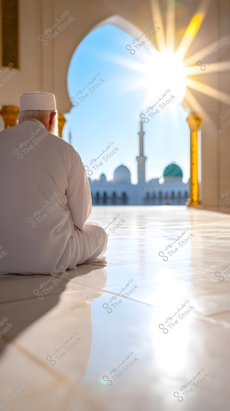 A man wearing a white robe and a white head covering is sitting inside a mosque. He is seen from the back, sitting on the shiny marble floor, facing a large archway that reveals a view of a mosque with minarets and domes. The sun is shining through the opening, creating beautiful light effects.