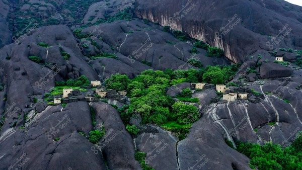 منظر جوي لقرية قديمة وسط جبال صخرية كبيرة في المملكة العربية السعودية. تظهر المباني الحجرية الصغيرة متناثرة بين الصخور الضخمة وعلى جوانب الجبال, وتحيط بها أشجار خضراء كثيفة تضفي على المكان لمسة طبيعية مميزة.